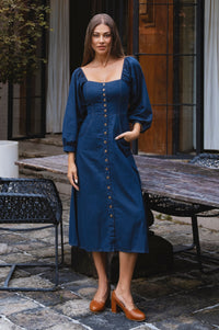 Woman wearing a blue linen dress standing outdoors on a stone patio.