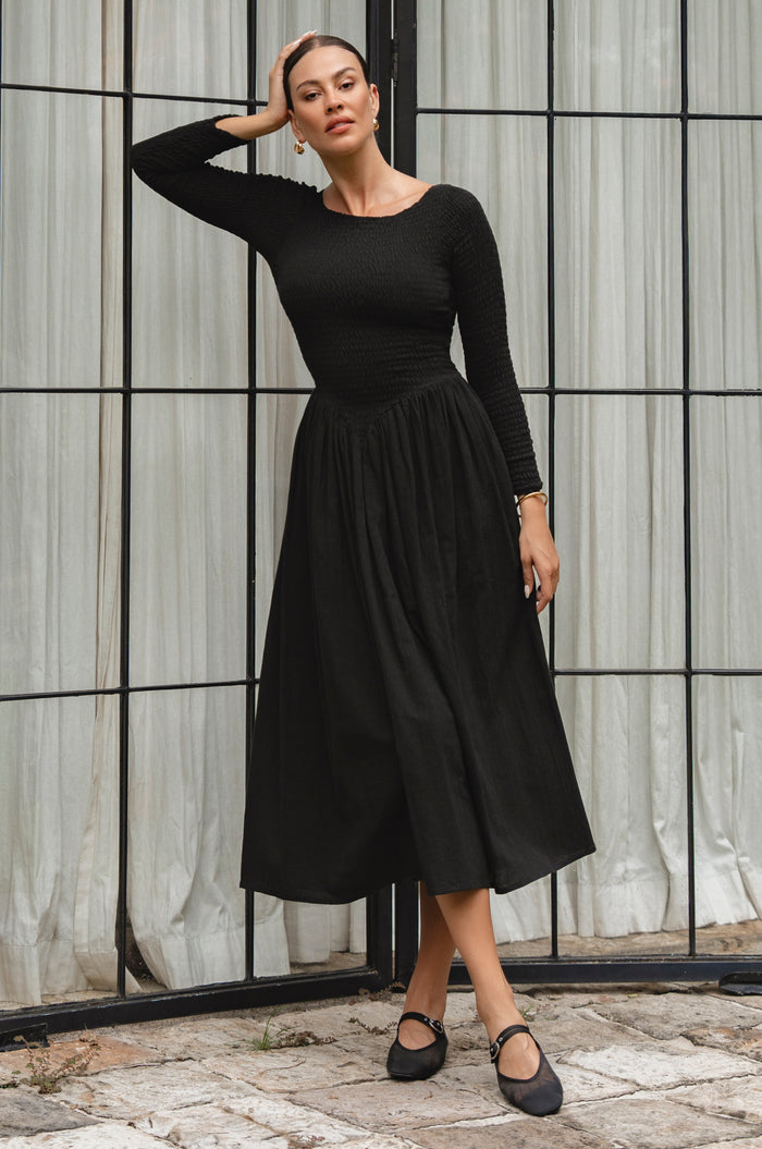 Woman wearing a black smocked dress standing against a textured wall.
