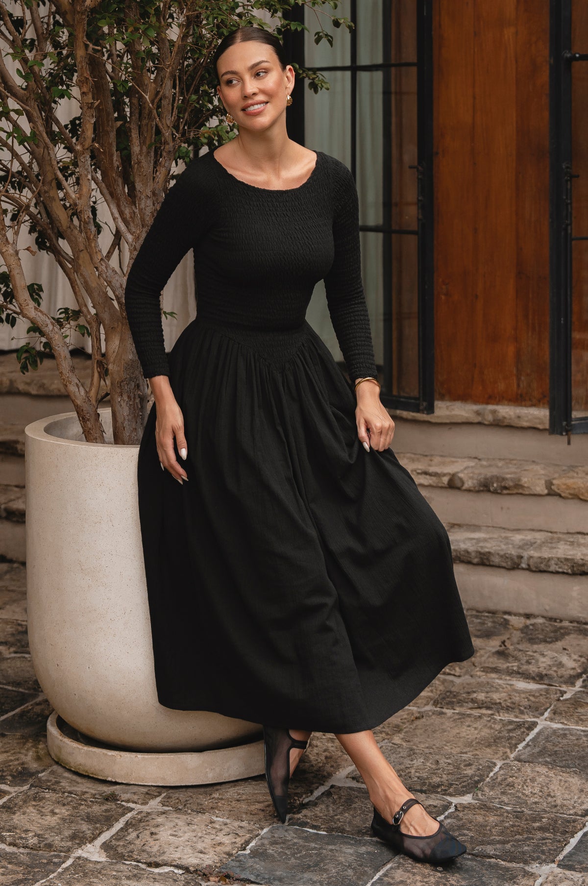 Woman wearing a black smocked dress standing outdoors on stone steps.