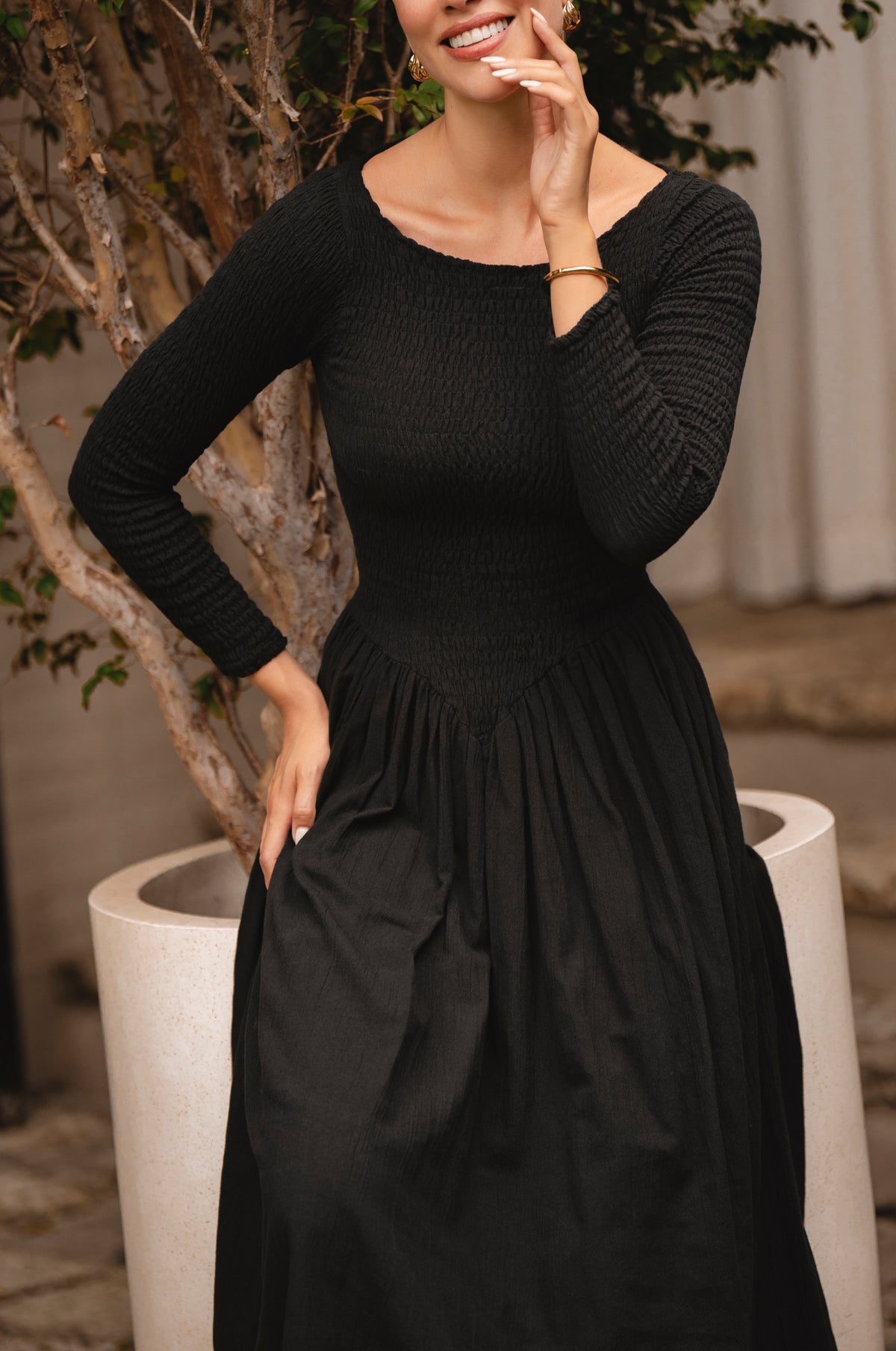 Woman wearing a black midi dress standing next to a decorative plant indoors.