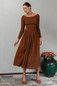 Woman wearing a brown midi linen dress standing against a gray wall with a decorative vase in the background.