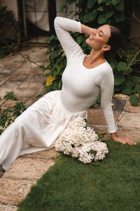 Woman in a white smocked long sleeve midi dress sitting on grass with flowers, surrounded by greenery.