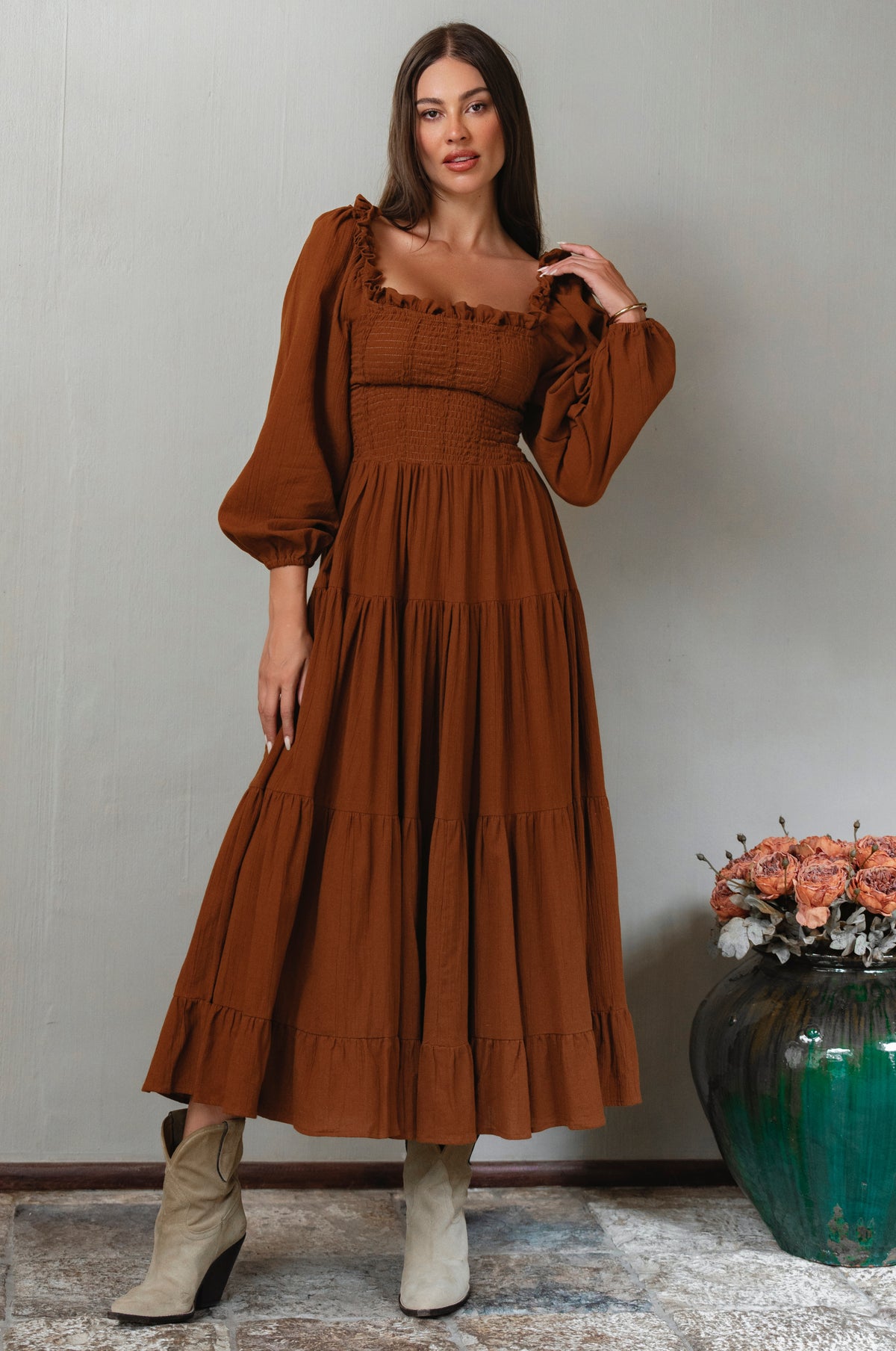 Woman wearing a brown dress with long sleeves standing against a plain wall with a vase of flowers in the background.