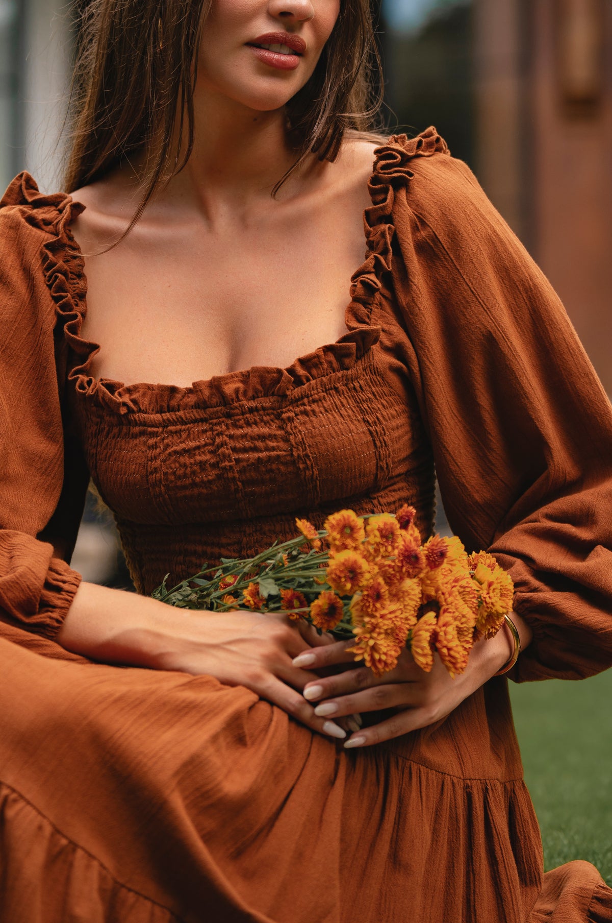 Woman in a smocked brown linen dress holding flowers with a blurred background