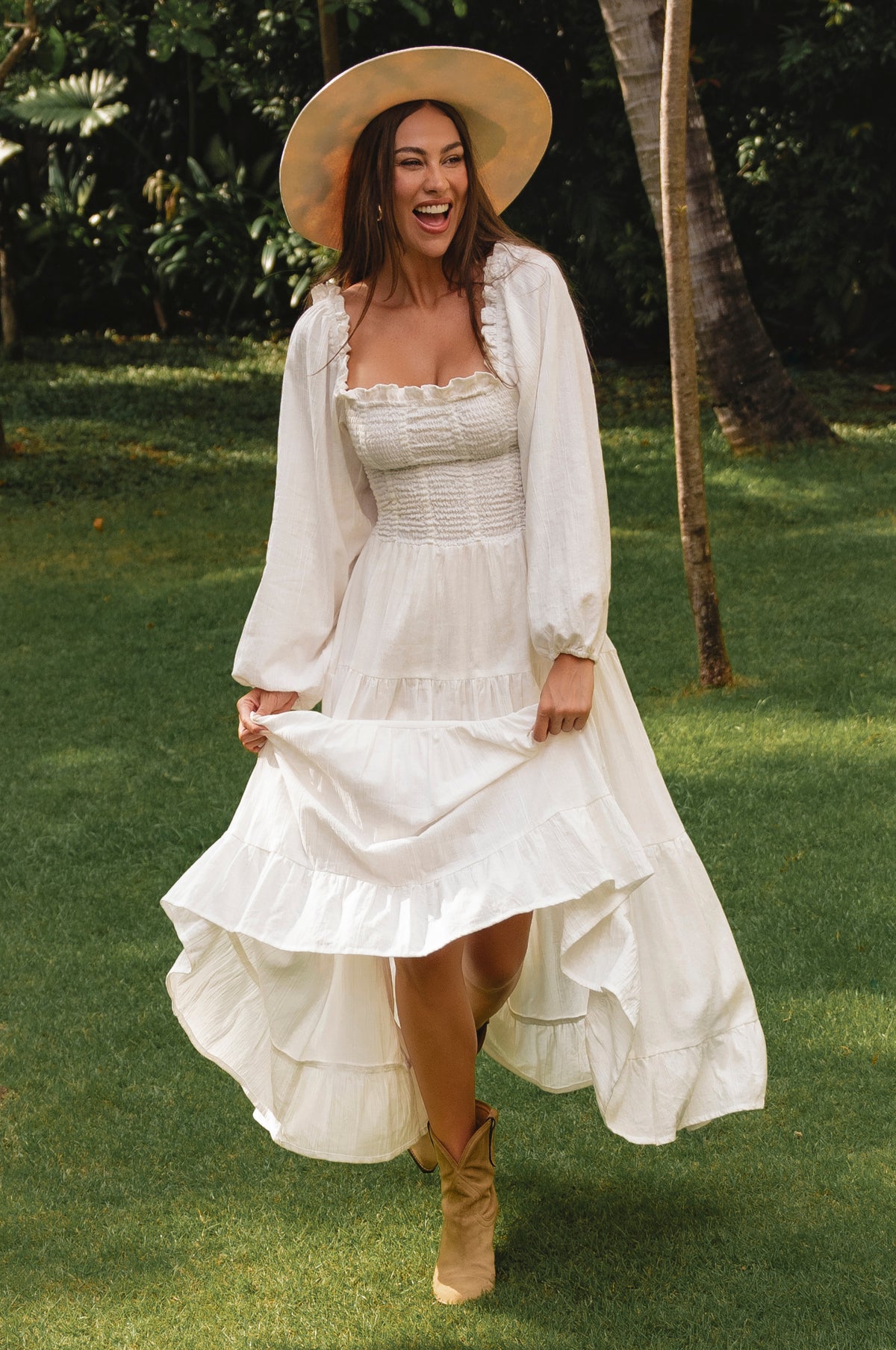 Woman in a white dress and hat standing on grass with trees in the background cottage core style