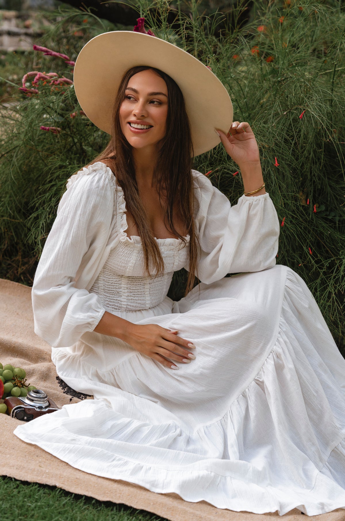 Woman in a white linen dress and beige hat sitting on a blanket outdoors cottage core style