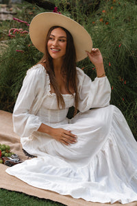 Woman in a white linen dress and beige hat sitting on a blanket outdoors cottage core style