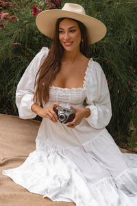 Woman in a white linen dress and hat holding a camera outdoors cottage core style