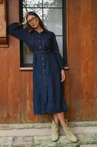 Woman in a nay denim dress and boots standing against a wooden wall.