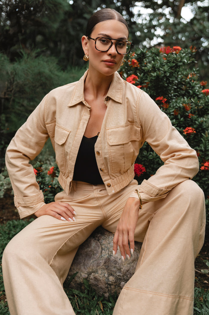 Woman in beige jacket and pants sitting outdoors with greenery and flowers in the background - cottage core style