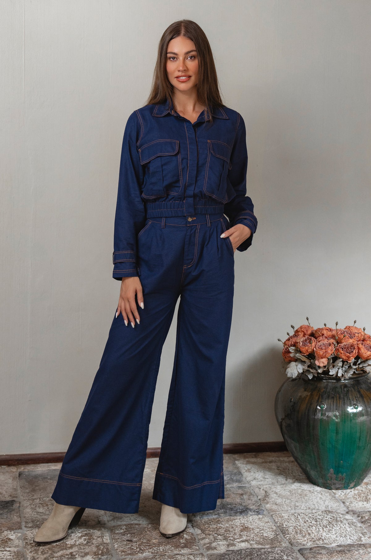 Woman wearing a blue denim set standing next to a vase with flowers against a plain wall - cottage core style