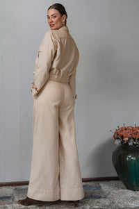 Woman wearing a beige outfit standing in a room with a vase of flowers on a table.
