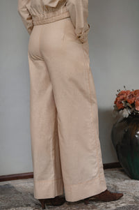 Woman wearing beige linen pants standing against a neutral background with a vase of flowers.