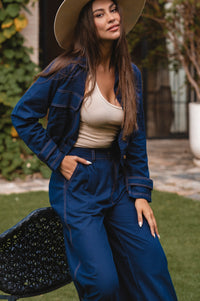 Woman in a navy linen denim like outfit with a beige hat standing outdoors.