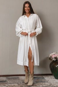 Woman wearing a white long sleeve dress with a belt, standing against a plain wall.