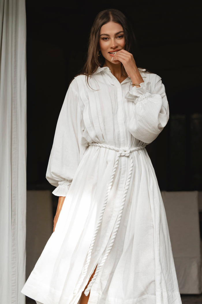 Woman wearing a white linen dress standing indoors with a dark background